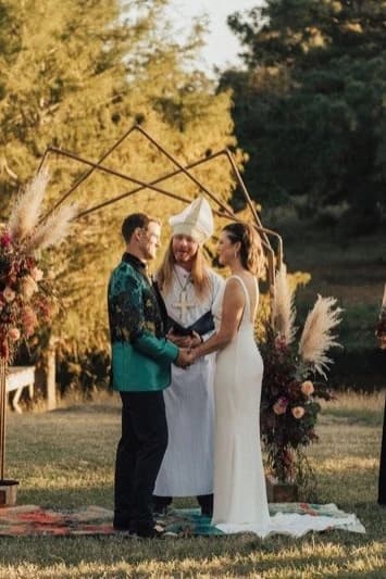A couple in wedding attire holding hands before an officiant at an outdoor golden-hour ceremony, beneath a geometric wooden arch flanked by pampas-grass and floral arrangements