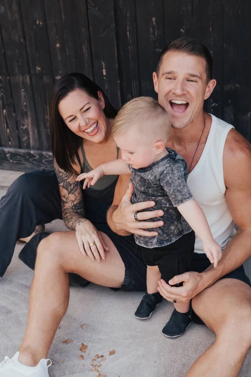 Three people laughing together while seated outdoors, one holding a young child